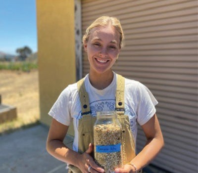 A student in overalls holds a large jar of grain outdoors near an agricultural building.