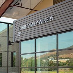 The exterior of the Lohr Family Winery building features modern signage and large windows with vineyard hills in the background.