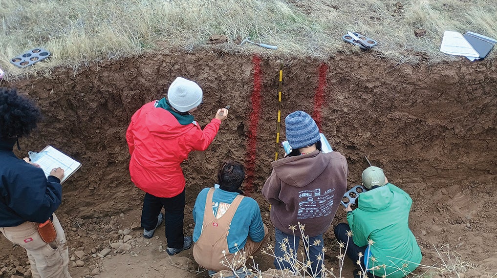 A group of five students stand in a hole in the ground judging the different layers of soil. 