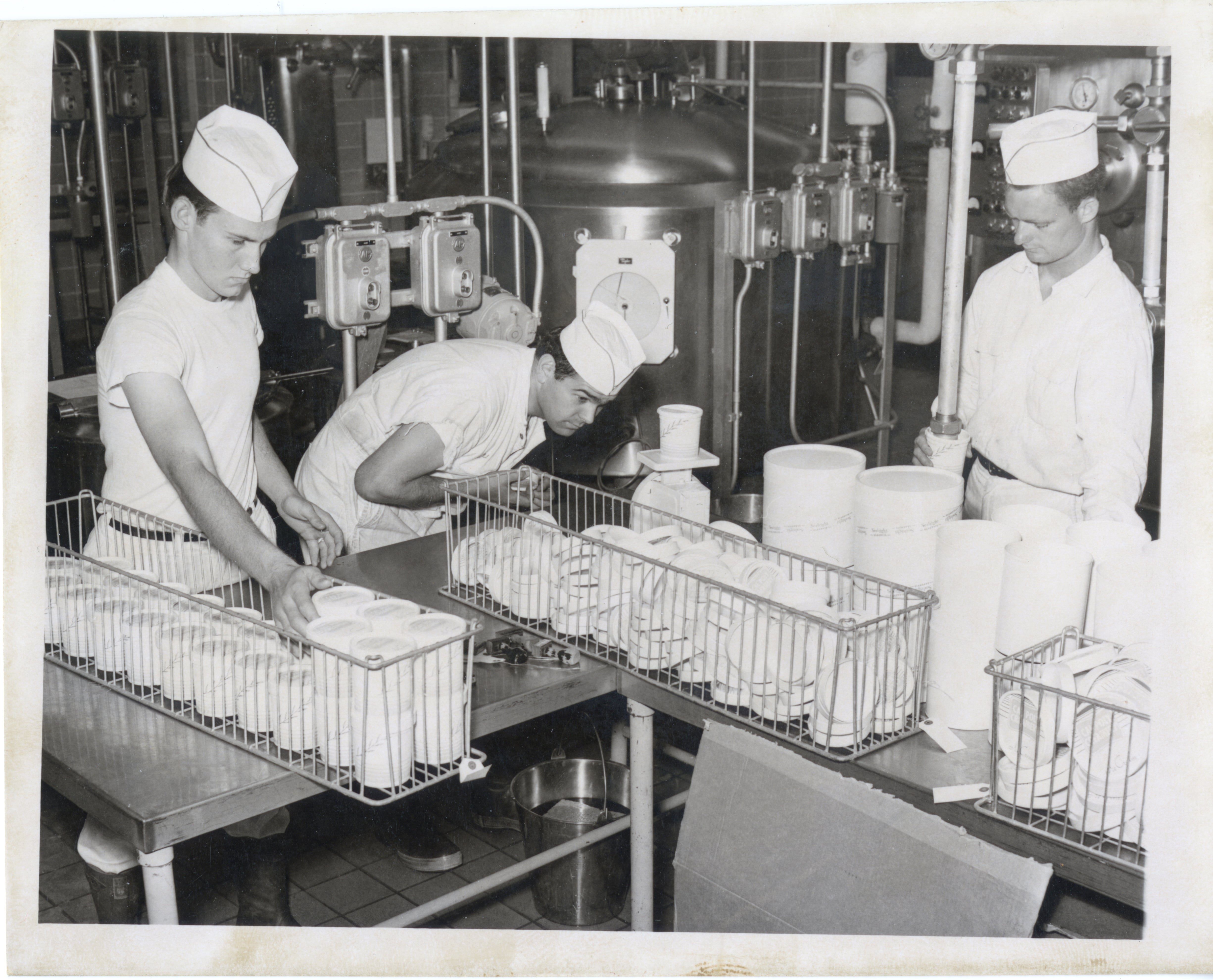 The students are reaching down into a freezer machine (a Cherry-Burrell Nu-Vat) to handle frozen food. They all wear the same uniform, including a white hat, circa 1950s.
