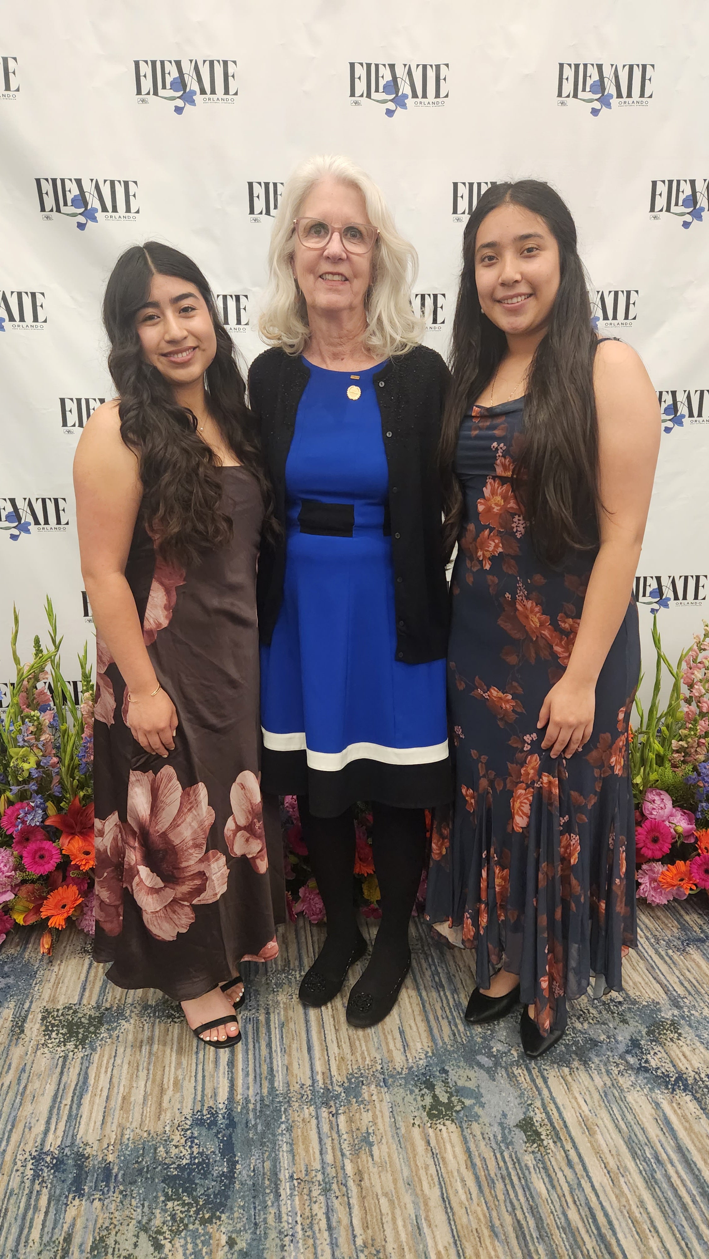 Two students wearing dresses stand next to their advisor. 