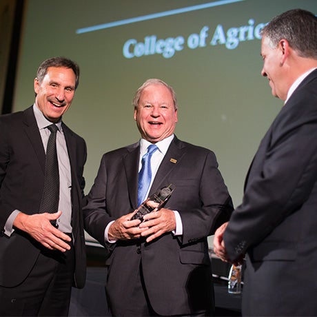 Three individuals in formal attire standing on stage during a College of Agriculture awards event, with one recipient holding a recognition award.