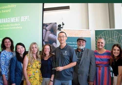 Group of faculty, staff, and students posing together at an awards ceremony, with one attendee holding a recognition plaque in front of presentation displays.