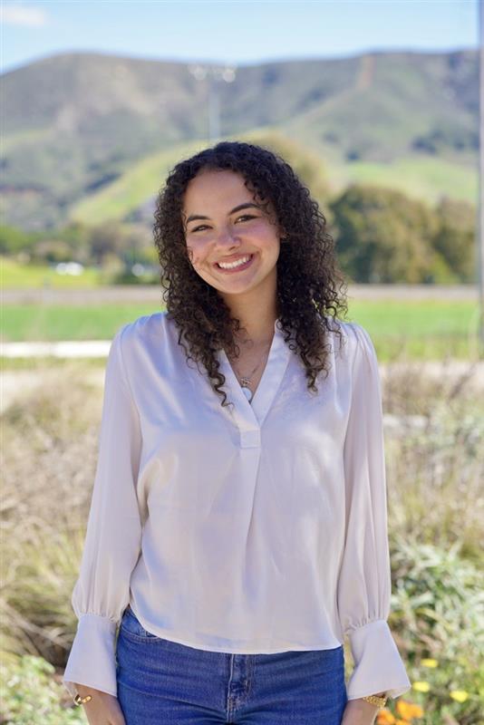 A smiling person stands outdoors in front of rolling hills and a natural landscape while wearing a light-colored blouse and jeans.