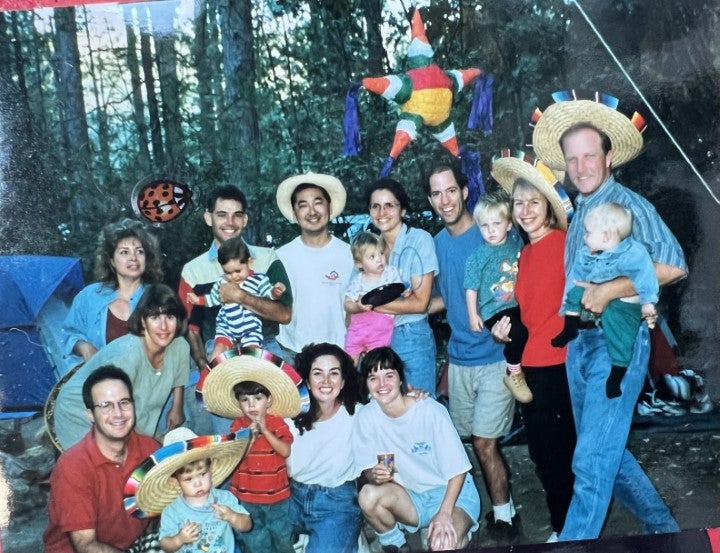 A large group of adults and children pose together at a campsite, some wearing festive sombreros, with tents and trees in the background.