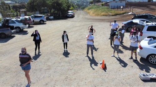 A group of people stand spaced apart in an outdoor lot while a drone flies overhead during a drone demonstration or training activity.
