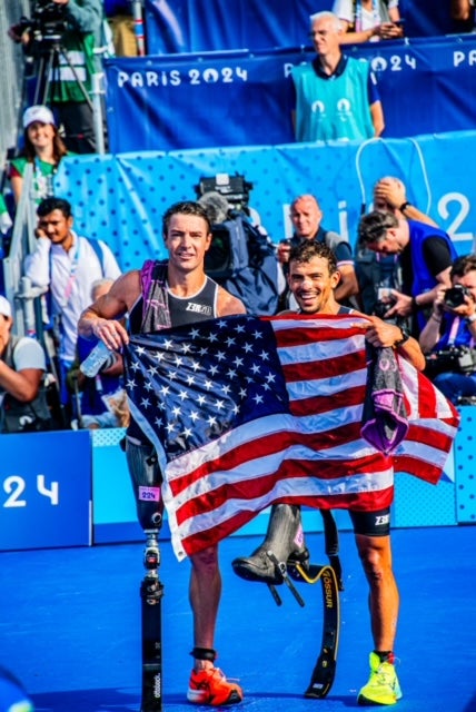 Two athletes wearing prosthetic running blades celebrate while holding American flags at a sporting event, with “Paris 2024” signage and photographers visible in the background.