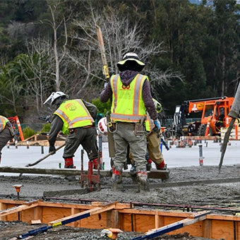 Construction crew pouring and leveling concrete foundation slabs while wearing high-visibility safety gear at a building site.