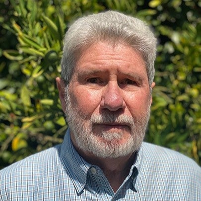 Professional outdoor portrait of a man with gray hair and beard standing in front of greenery.
