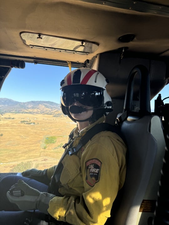 A CAL FIRE crew member wearing a flight helmet and protective gear sits inside a helicopter during an aerial wildfire or emergency response operation.