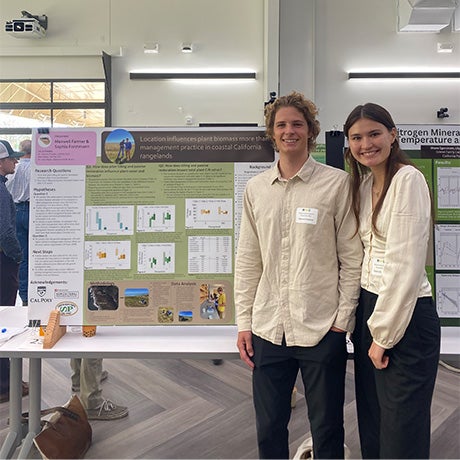 Two students stand beside a research presentation poster during an academic showcase or symposium in a modern indoor event space.