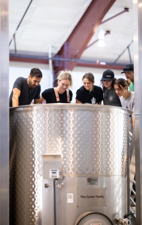 A group of people gathers around a large stainless steel fermentation tank while observing the winemaking process.