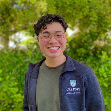 A smiling person wearing glasses and a Cal Poly jacket stands outdoors with greenery in the background.