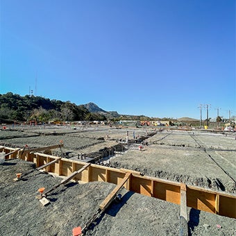 Wide view of a large building under construction with steel framing, roofing installation, heavy equipment, and construction materials spread across the site.