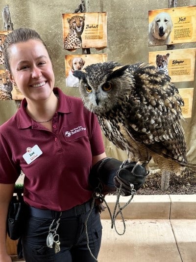 Wildlife educator holding a large owl during an animal education or conservation outreach presentation, with informational displays about animals in the background.