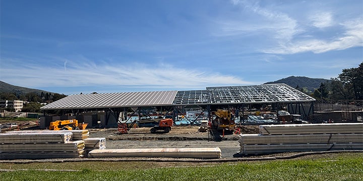 Early-stage construction site showing concrete foundations, framing outlines, and excavation work beneath a clear blue sky with hills in the background.