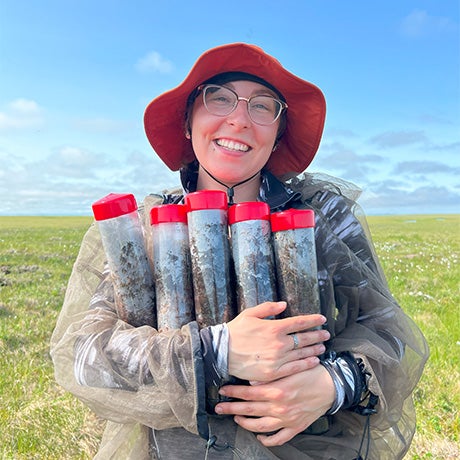 Researcher smiling while holding multiple soil sample tubes during outdoor environmental or agricultural fieldwork in a grassy landscape.