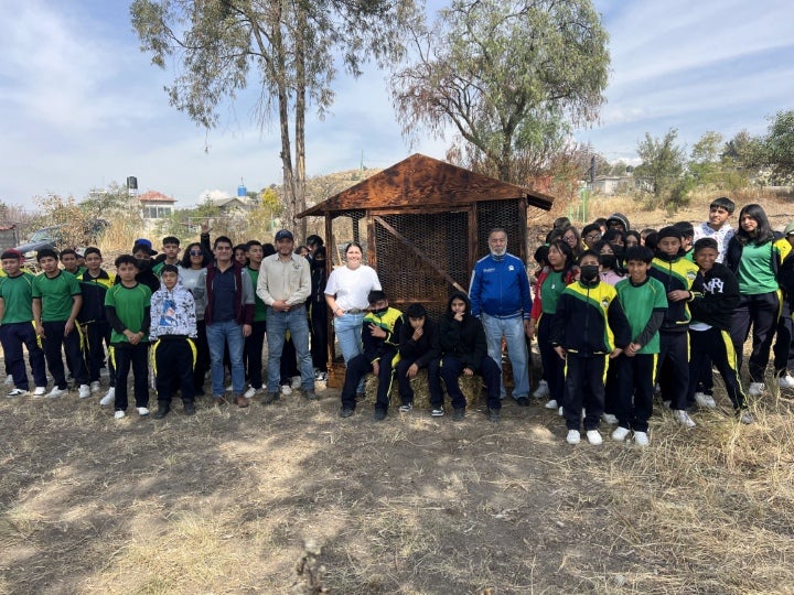 A large group of students and adults stands outdoors around a small wooden structure in a dry grassy landscape with trees in the background.