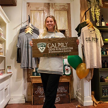 Student standing inside a campus merchandise or alumni store holding a College of Agriculture and Environmental Sciences sign, surrounded by Cal Poly apparel and promotional items.