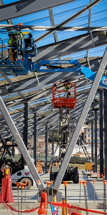 Construction workers operating elevated lifts while assembling the steel framework of a large building structure at an active construction site.