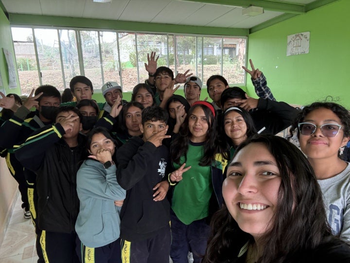 A group of students gathers indoors for a group selfie, smiling and posing together inside a classroom with green walls.