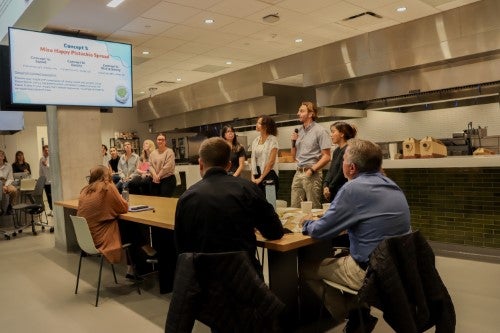 A group of presenters speaks to an audience in a large kitchen-style event space while attendees sit at tables listening to the discussion and viewing a presentation screen.
