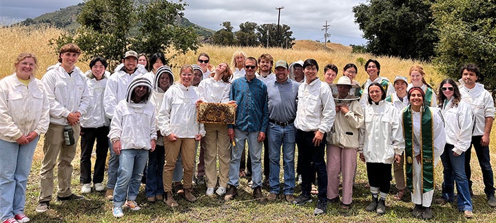 A large group of people wearing protective beekeeping jackets gathers outdoors in a grassy field, with some holding honeycomb frames and beekeeping equipment.