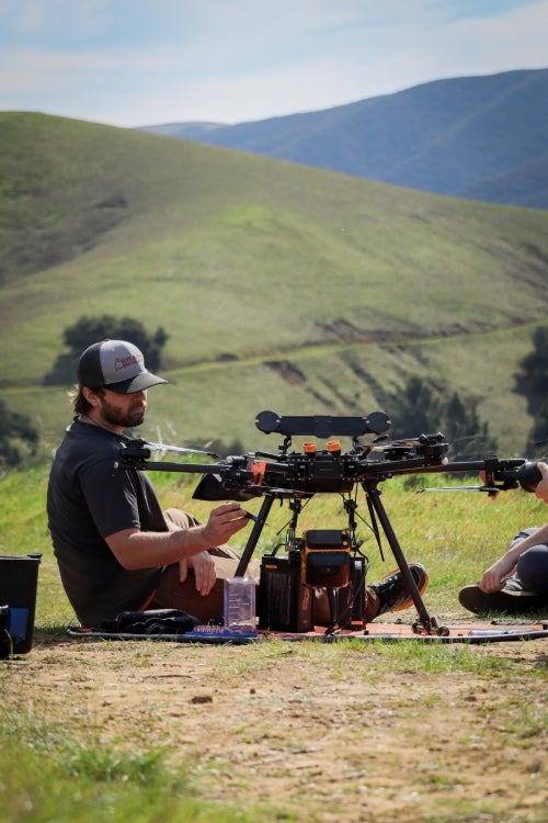 A researcher adjusts a large drone in an open field surrounded by rolling hills.