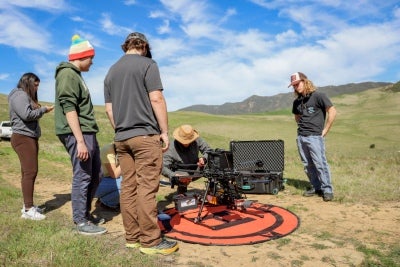 A group of students and researchers gather around a large drone during an outdoor fieldwork or technology demonstration in a grassy landscape.