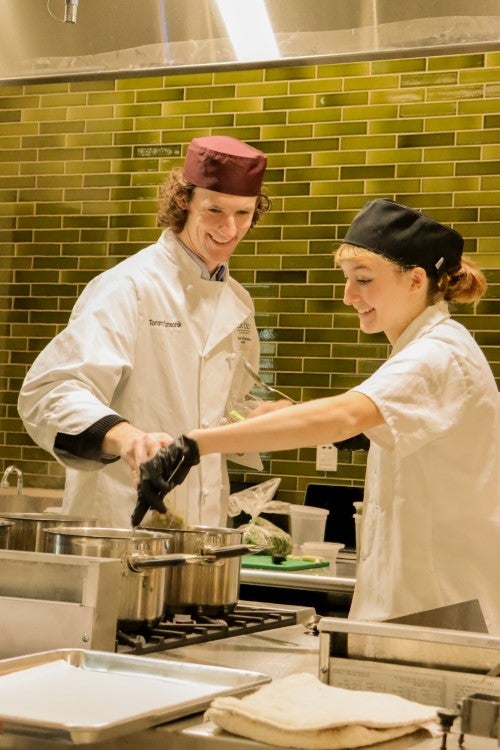 Two culinary students wearing chef uniforms and hats smile while cooking together at a stovetop in a commercial kitchen.
