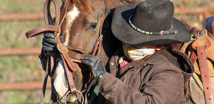 Close-up of a person in western riding attire adjusting tack on a horse while standing beside it.