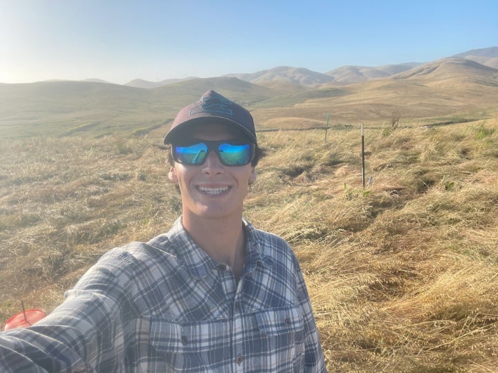 Person wearing sunglasses and outdoor workwear taking a selfie in a dry grassland landscape with rolling hills and open countryside in the background.