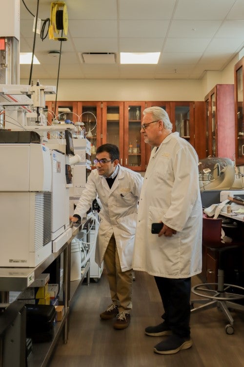 Two researchers in white lab coats working together in a laboratory, examining scientific equipment and instrumentation in a research facility with cabinets and analytical machines surrounding them.