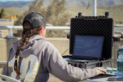 A person works on a laptop connected to field equipment outdoors during a drone or mapping operation.