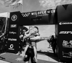 Two people celebrate with a hug near the finish line at an outdoor endurance or cycling event, with event signage visible in the background.