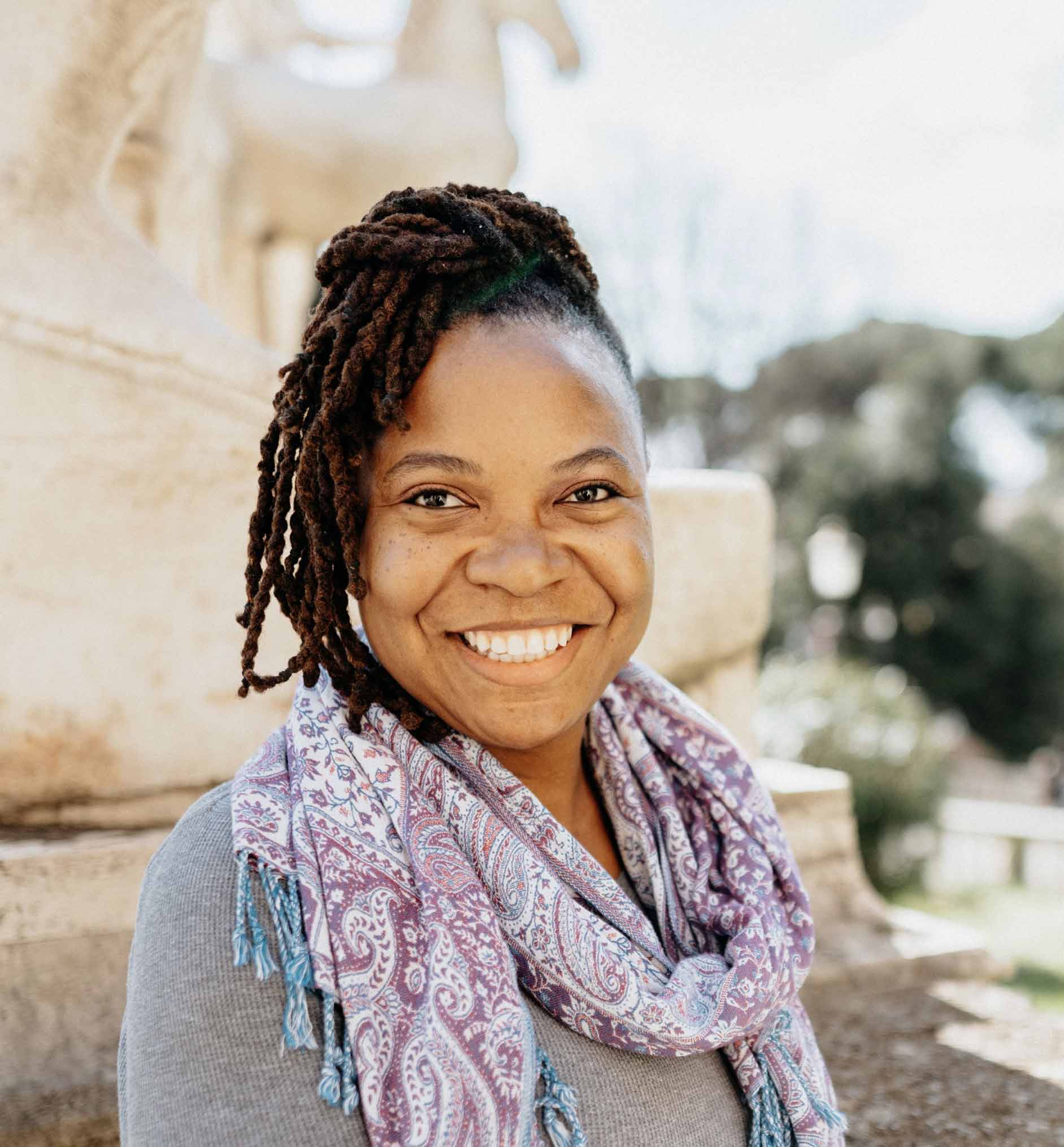 Portrait of a smiling woman with braided hair wearing a patterned scarf and gray sweater while standing outdoors near stone architecture.