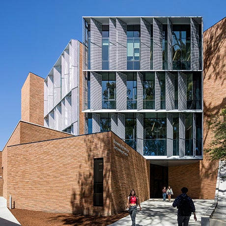 People walk near a modern multi-story academic building featuring brick walls, large glass windows, and geometric metal shading panels.