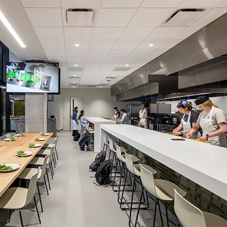 People prepare food in a modern commercial kitchen with long counters, bar seating, and a dining area set with plated dishes.