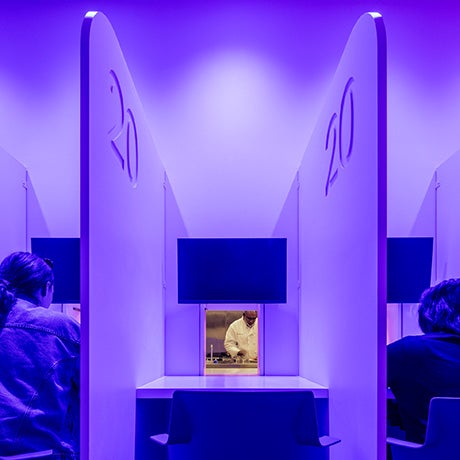 Students sit in individual booths inside a culinary lab, each station featuring a viewing window into the kitchen where cooking demonstrations and food preparation can be observed.