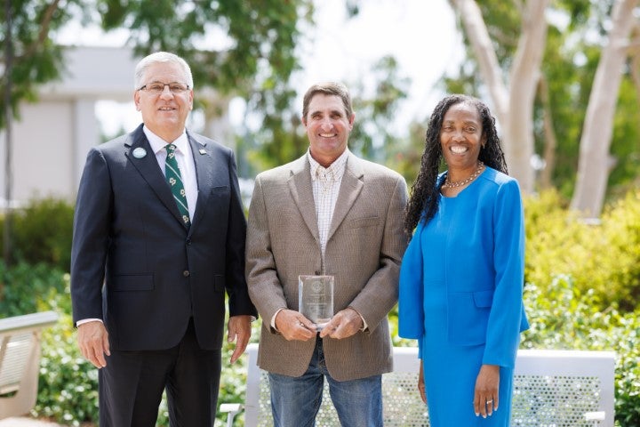Three individuals posing outdoors during an award recognition event, with one person holding an achievement plaque.