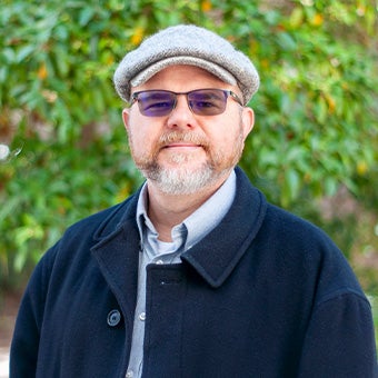 Portrait of a man with glasses, a gray flat cap, and a trimmed beard standing outdoors in front of green foliage while wearing a dark coat and collared shirt.