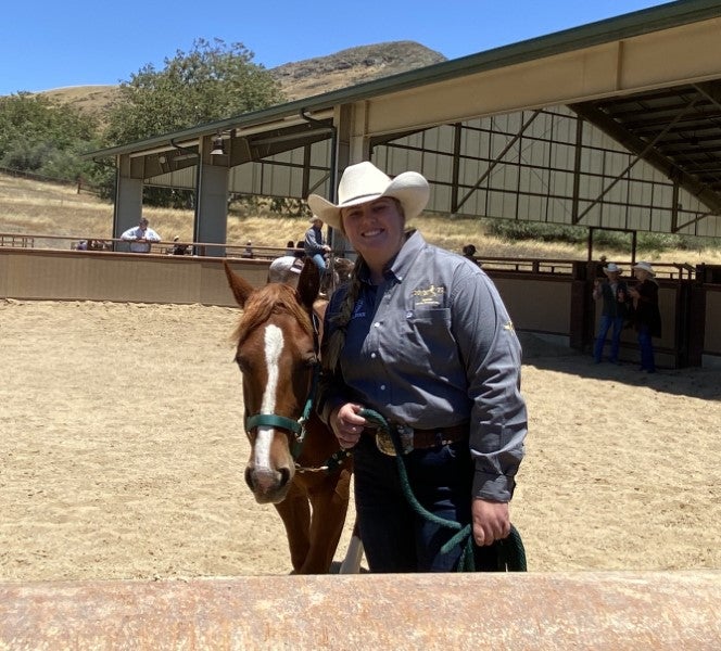 Woman in western attire and cowboy hat standing beside a horse in an outdoor equestrian arena at a livestock or horse event.