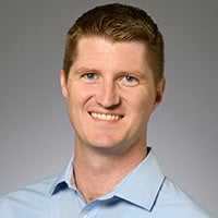 Professional headshot of a man wearing a light blue collared shirt against a neutral studio background.