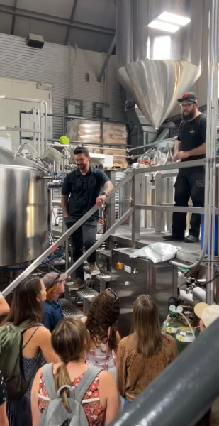 Two instructors stand on an industrial brewing platform while speaking to a group of students during a brewery or fermentation science demonstration.