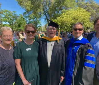 Group photo of faculty, graduates, and guests posing together outdoors during a university commencement celebration, with two individuals in academic regalia.