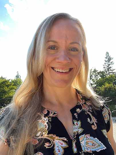 Portrait of a smiling woman with long blonde hair standing outdoors in natural sunlight wearing a patterned blouse.