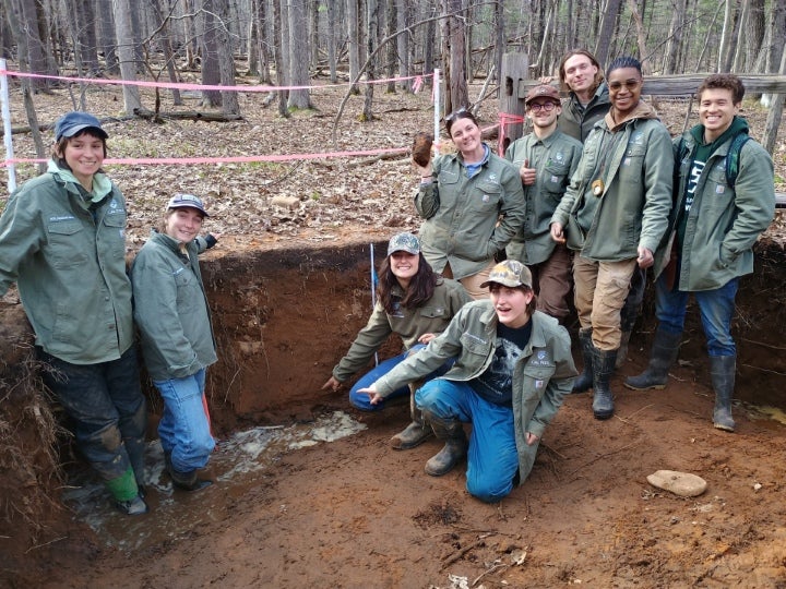 Students gathered around a soil excavation pit during a hands-on soil science or environmental field study activity in a forested area.