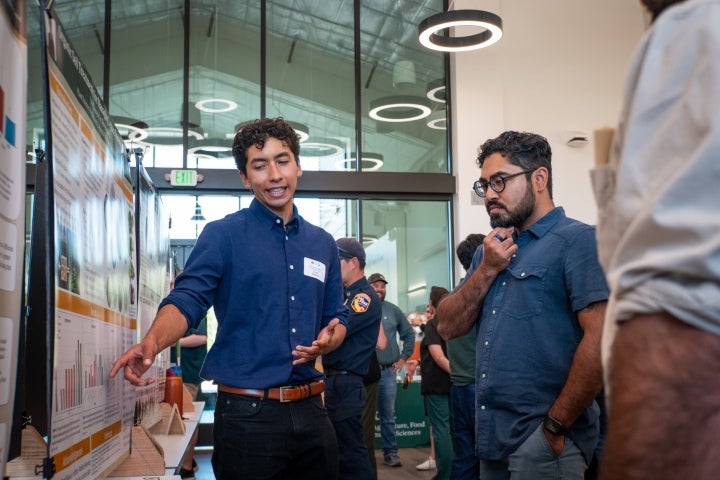 Student presenting research findings beside a scientific poster during a research symposium or academic showcase event.