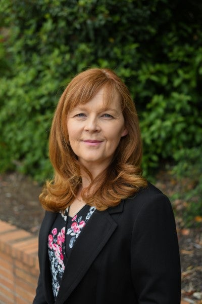 Professional portrait of a woman with red hair wearing a black blazer and floral blouse, standing outdoors in front of green foliage.