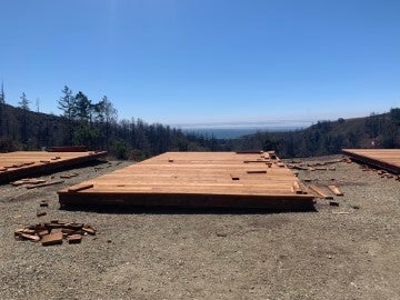Large wooden platform structures laid out on a cleared hillside construction site surrounded by trees and open sky, with building materials arranged across the landscape.